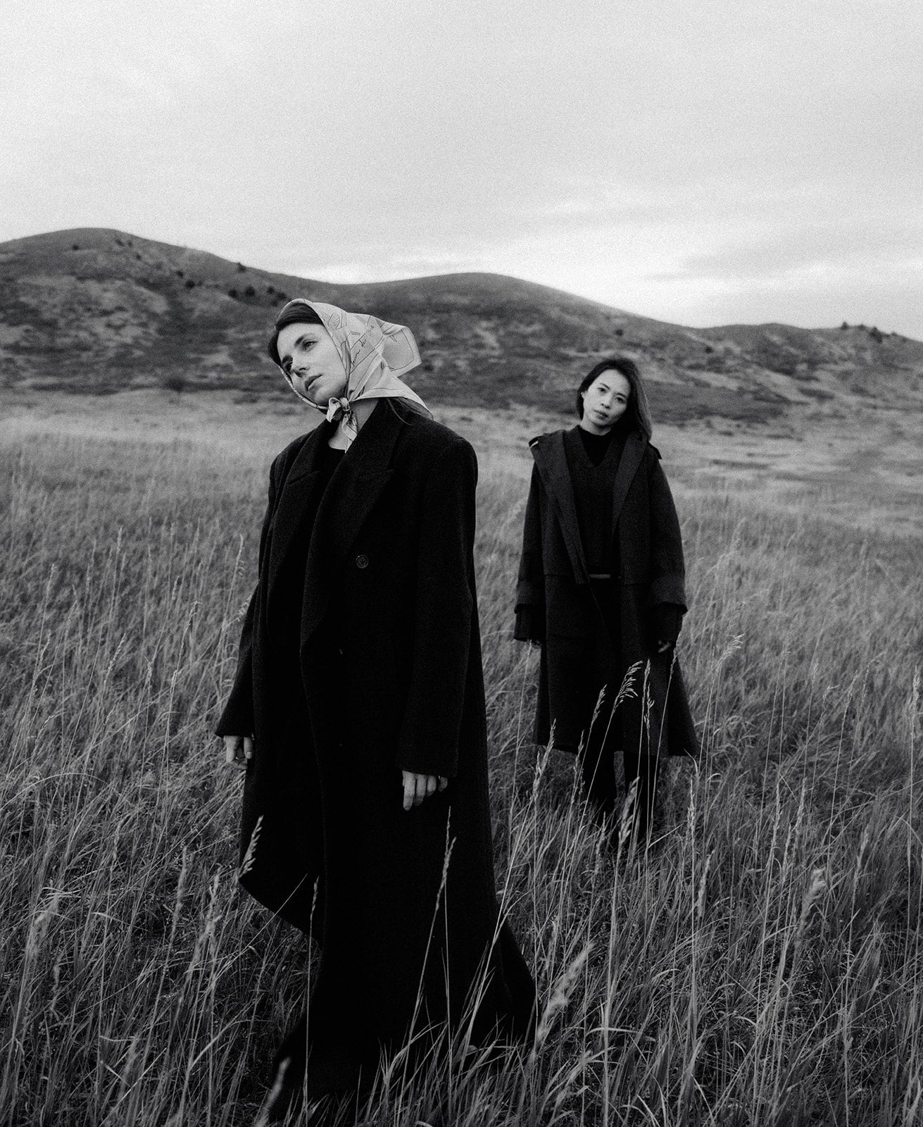 Two people in dark clothing standing in a field with hills in the background. Photoshoot for Skyler and Dasha. Shaa.Project