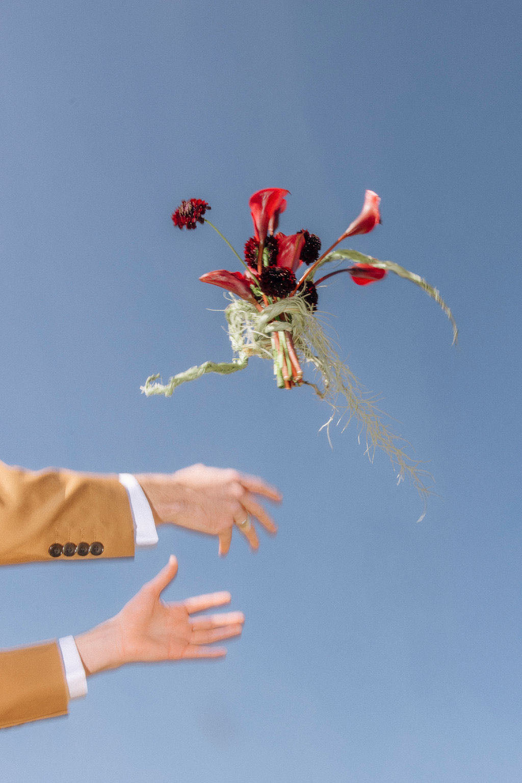 Bouquet of bridal flowers being thrown with a person's hands reaching out against a clear blue sky.