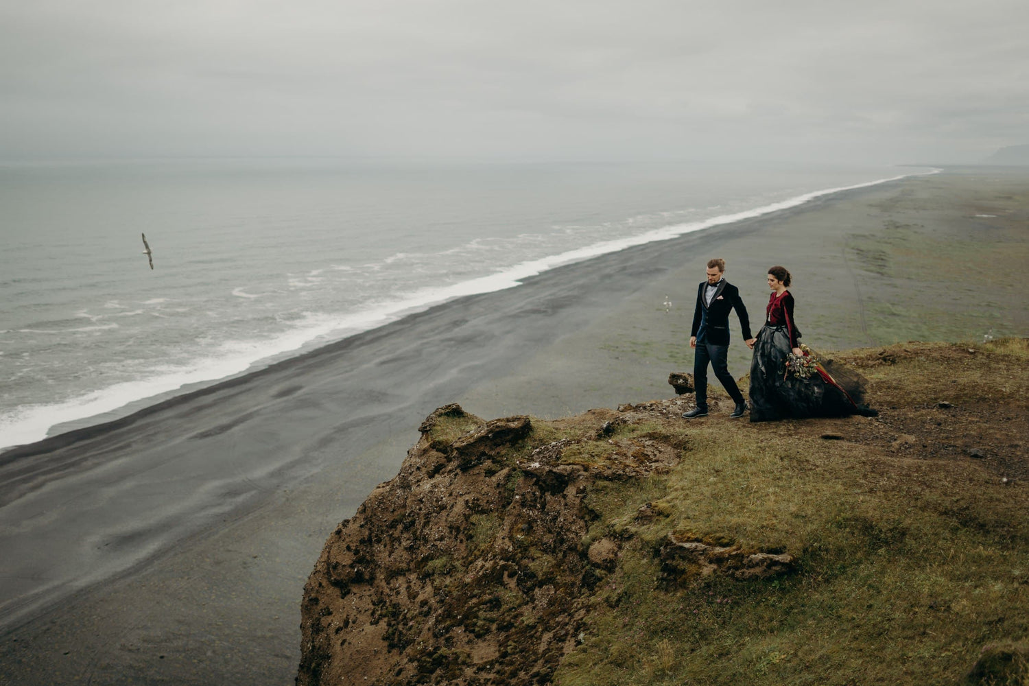 Destination wedding couple walking along coastal cliff with dramatic landscape and intimate bridal bouquet