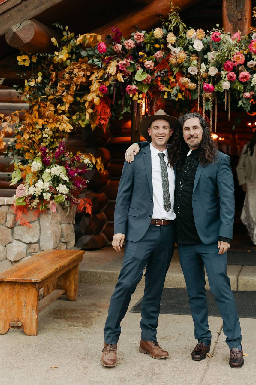 Two men in suits standing in front of a floral archway.